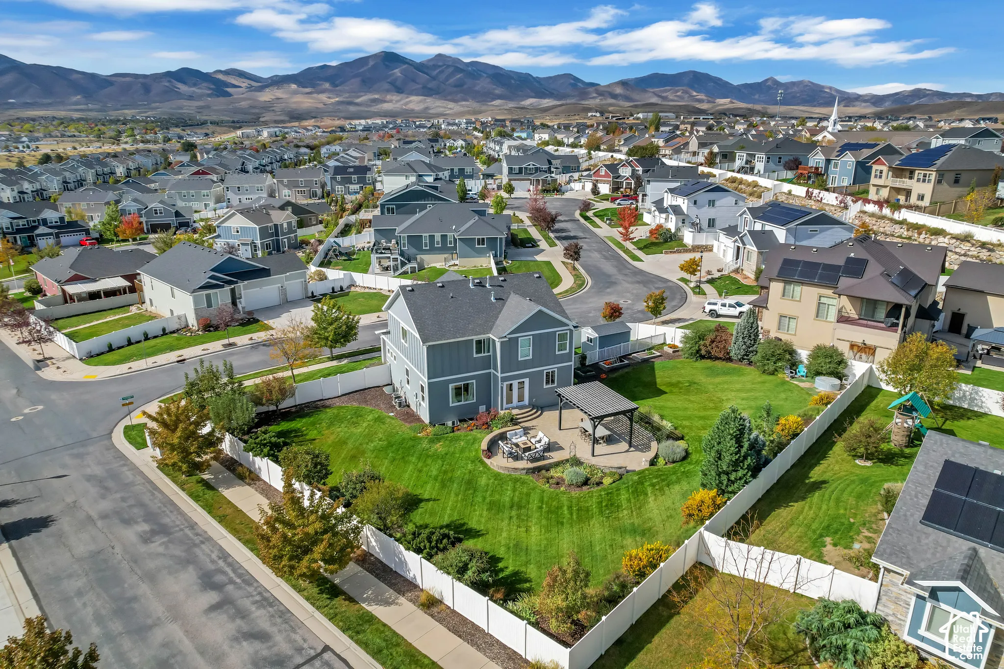 Corner lot property featuring a manicured lawn, stone paver patio, permanent pergola, and perimeter white vinyl fencing with a mountain backdrop