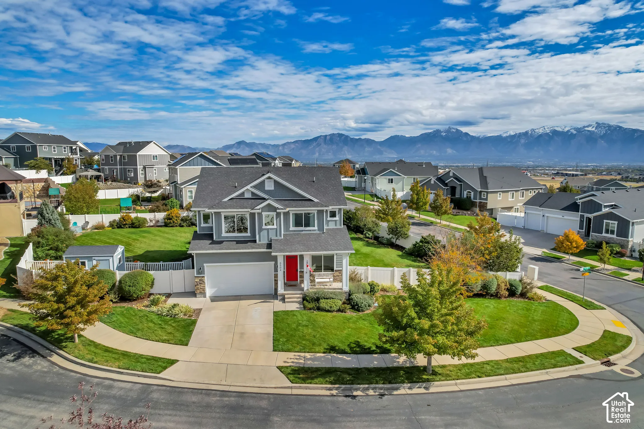 Two-story residential exterior featuring gray siding, stone veneer accents, a double-car garage, a white vinyl perimeter fence, and a corner lot position with mountain scenery
