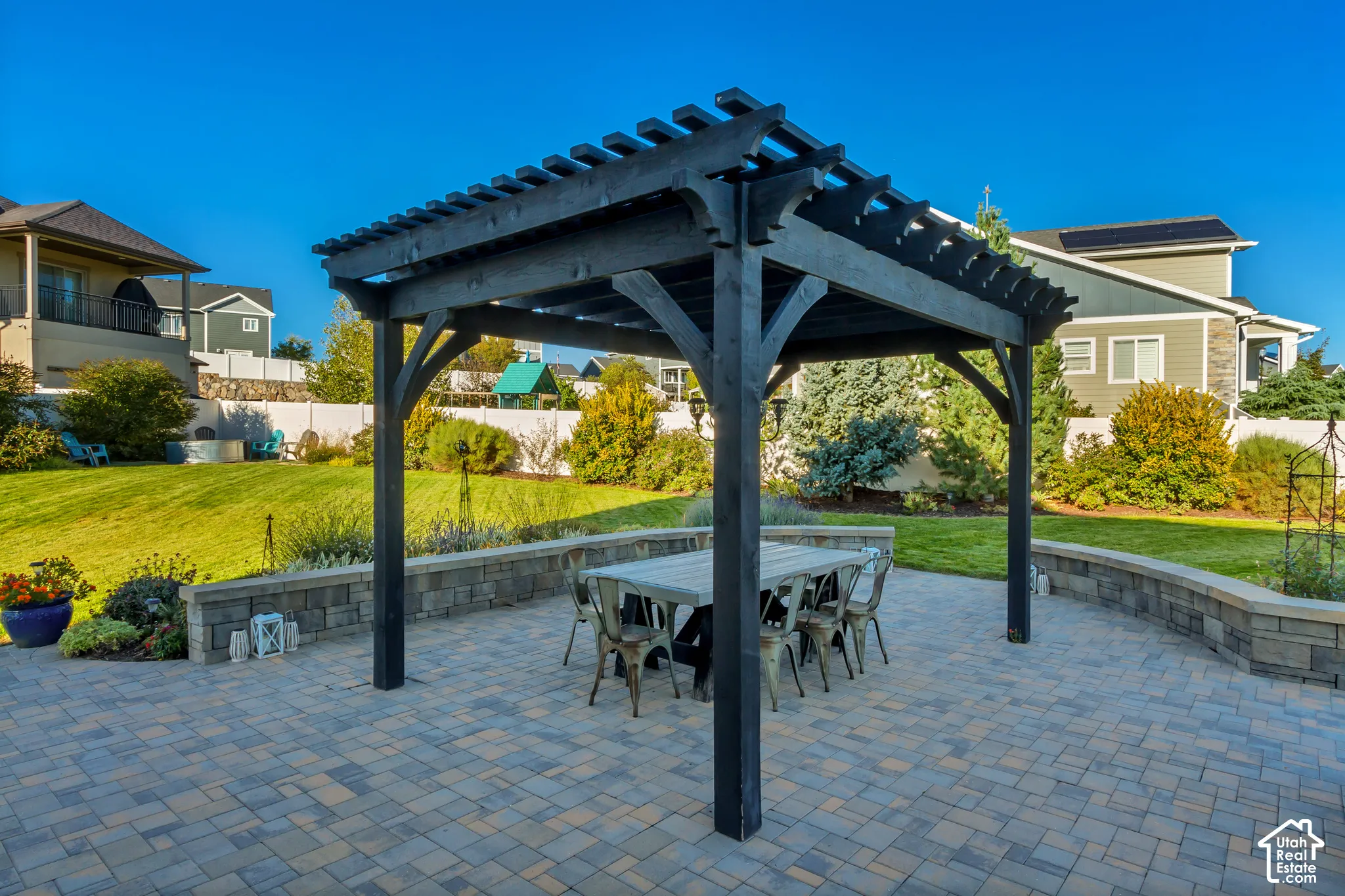 Backyard patio featuring pavers, a Western Timberframe wooden pergola, and a curved stone retaining wall surrounded by beautifully planted garden beds and an expansive grass lawn