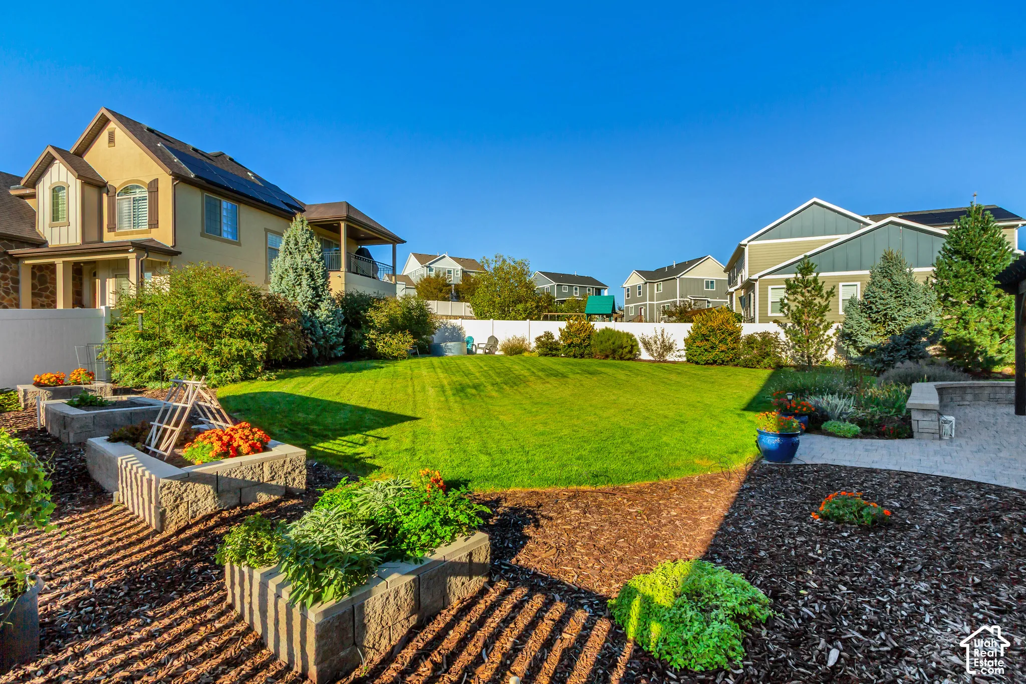 Backyard patio featuring pavers, a Western Timberframe wooden pergola, and a curved stone retaining wall surrounded by beautifully planted garden beds and an expansive grass lawnstone-block raised garden beds, wood-mulch landscaping, and perimeter vinyl f