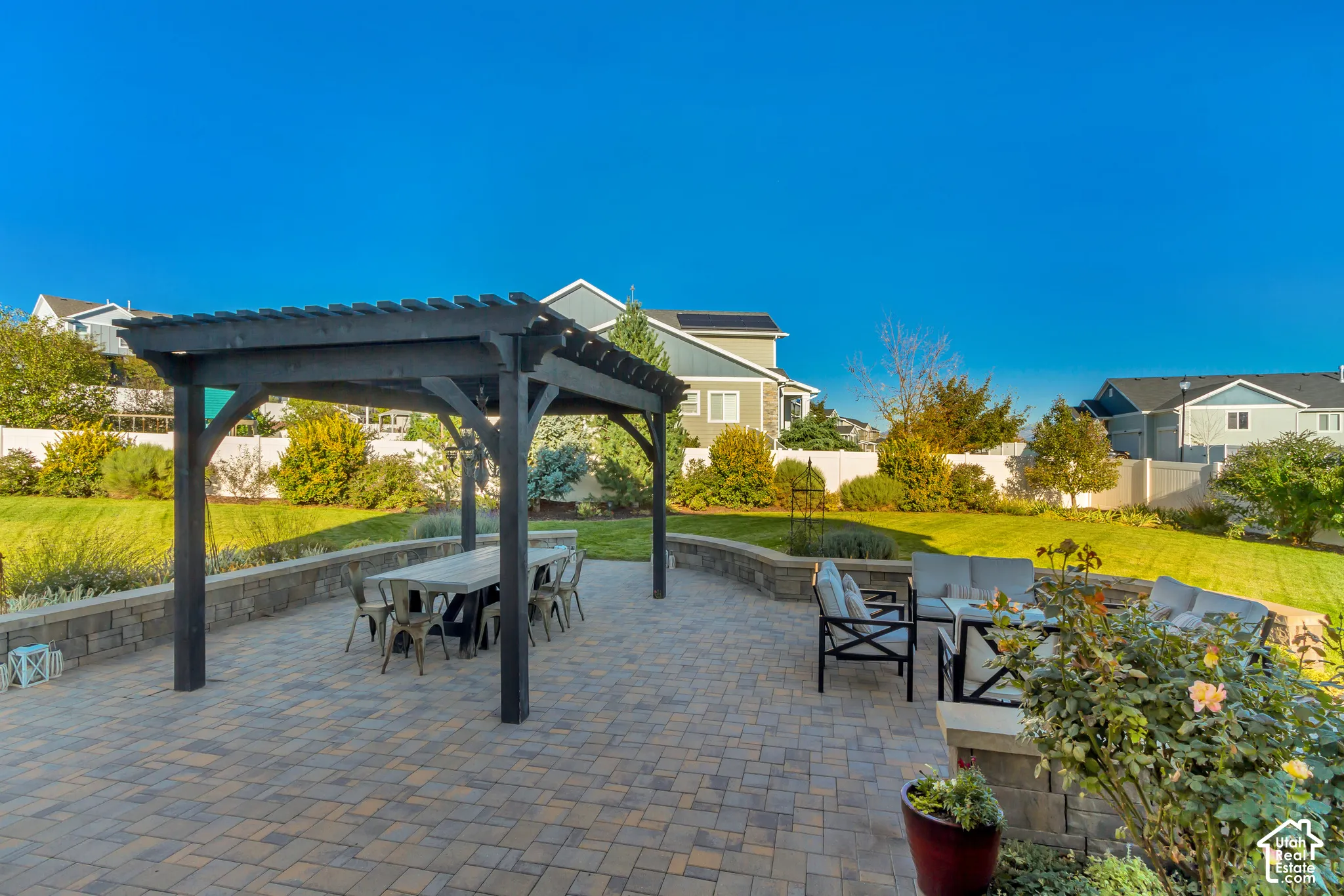 Backyard patio featuring pavers, a Western Timberframe wooden pergola, and a curved stone retaining wall surrounded by beautifully planted garden beds and an expansive grass lawn