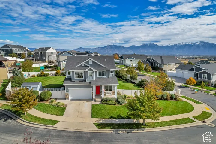 Two-story residential exterior featuring gray siding, stone veneer accents, a double-car garage, a white vinyl perimeter fence, and a corner lot position with mountain scenery