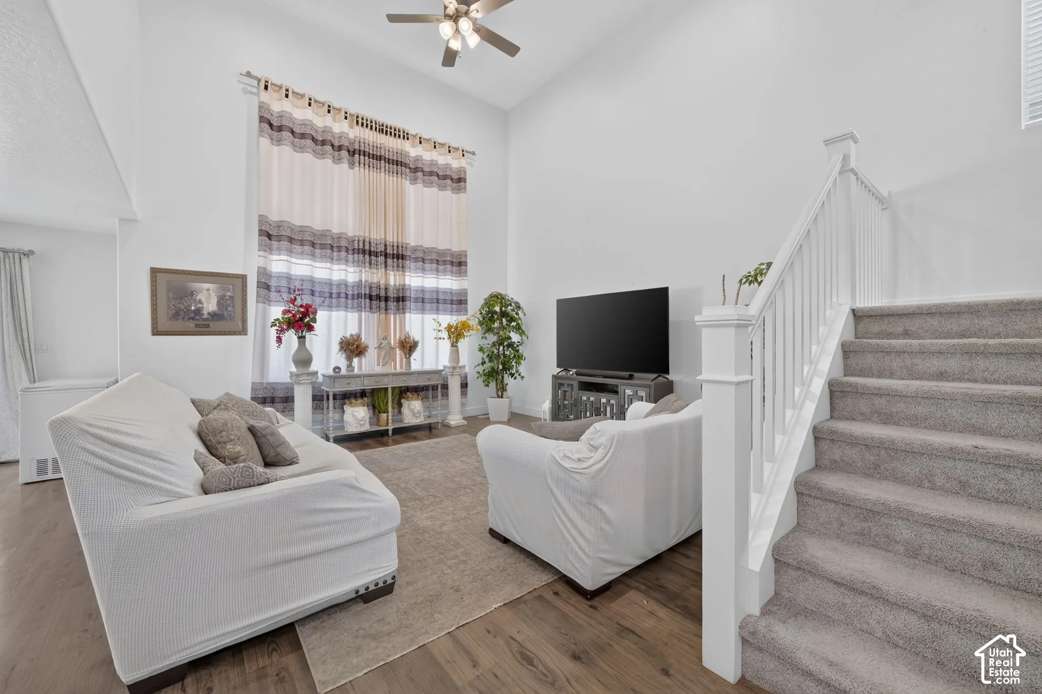 Bright open-concept living area featuring hardwood-finish flooring, a ceiling fan with integrated lighting, and abundant natural light from multi-pane windows dressed with striped drapery