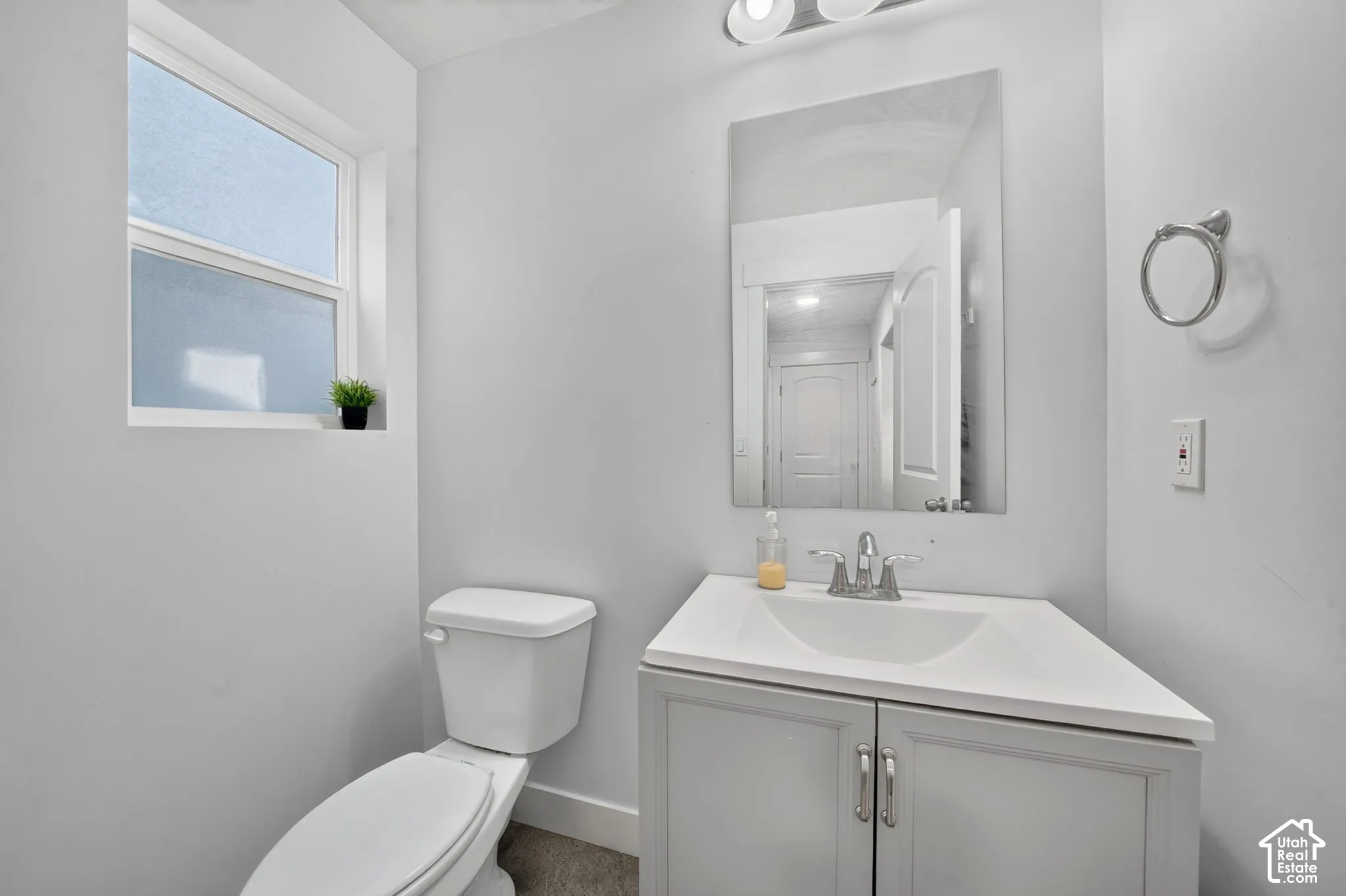 Clean and bright bathroom featuring a white vanity with dual-handle faucet and ample countertop space, a ceramic toilet, framed mirror with integrated lighting, and a large window providing natural light