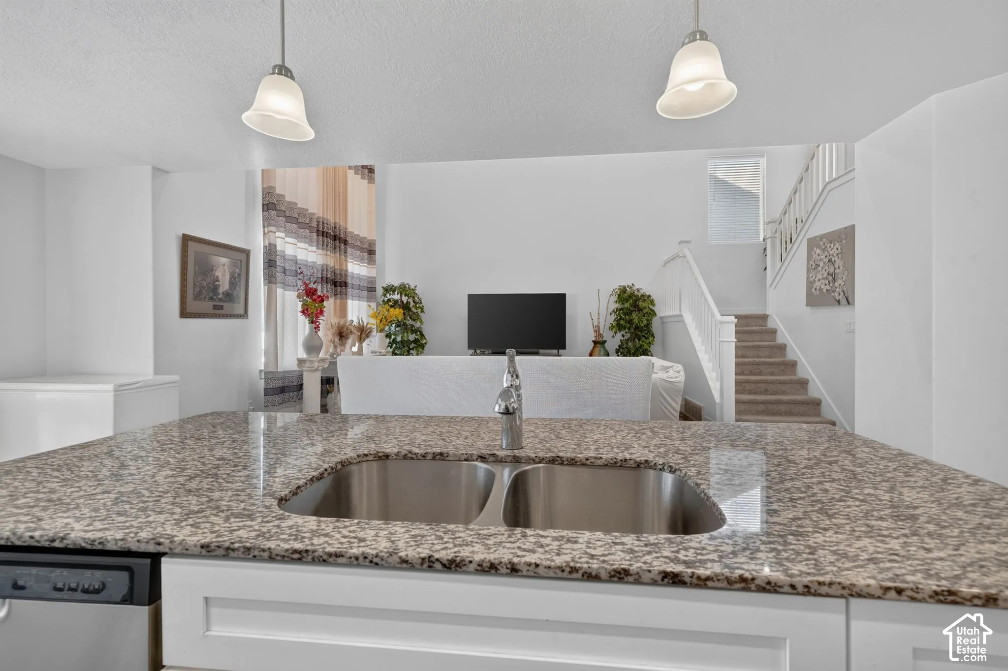 Open-concept kitchen and living area featuring a granite-topped island with dual undermount sinks, stainless steel appliances, pendant lighting, and white cabinetry