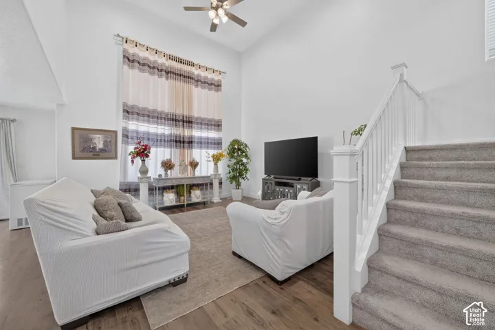 Bright open-concept living area featuring hardwood-finish flooring, a ceiling fan with integrated lighting, and abundant natural light from multi-pane windows dressed with striped drapery