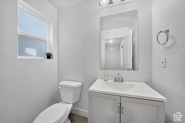 Clean and bright bathroom featuring a white vanity with dual-handle faucet and ample countertop space, a ceramic toilet, framed mirror with integrated lighting, and a large window providing natural light