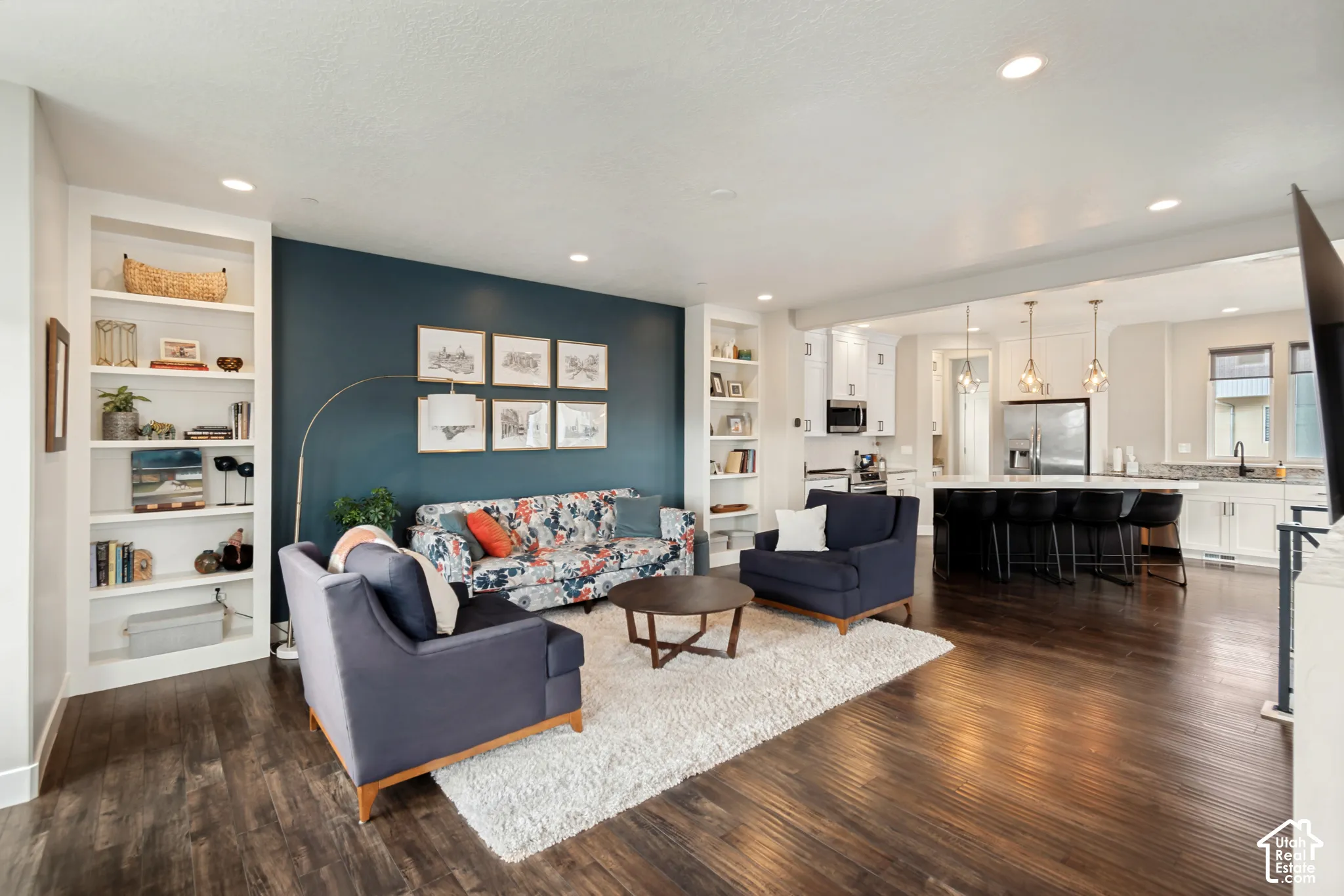 Wood-finish flooring throughout, living area with built-in shelving, kitchen with white cabinetry, central island with seating, and recessed lighting