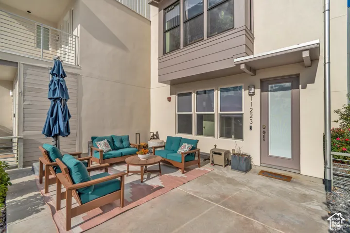 Paved patio area with a modern entry door, multiple windows, and a second-story balcony with metal railing