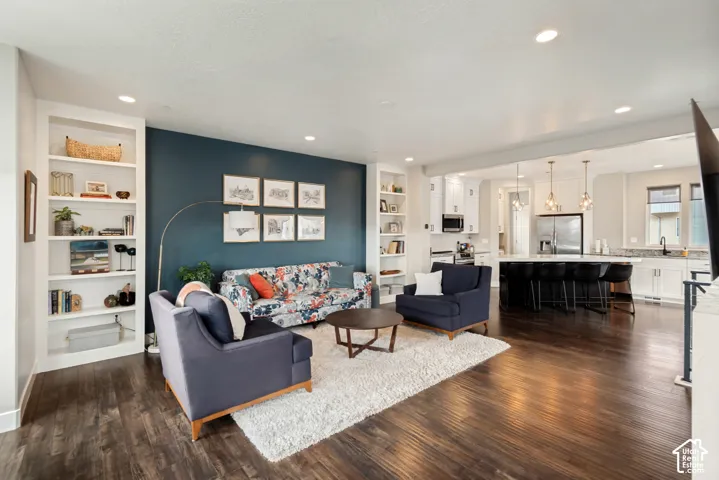 Wood-finish flooring throughout, living area with built-in shelving, kitchen with white cabinetry, central island with seating, and recessed lighting