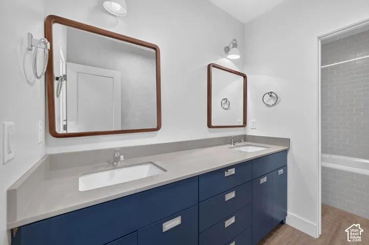 Dual undermount sinks with chrome fixtures, a long blue vanity, wood-finish flooring, subway tile tub surround, and two mirrors with wood frames