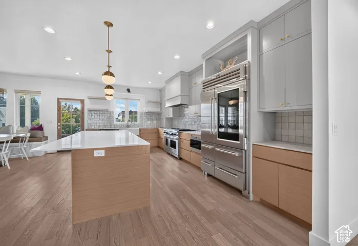 Spacious kitchen featuring a large central island with a solid surface countertop, light wood-finish flooring, integrated stainless steel appliances, light gray upper cabinetry, and light wood-finish lower cabinetry