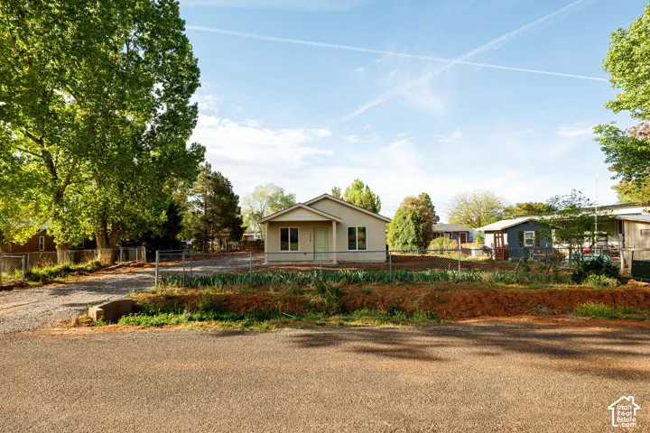 Single-story residence featuring light-toned siding, a covered front entryway, metal chain-link fencing, a gravel driveway, and mature trees