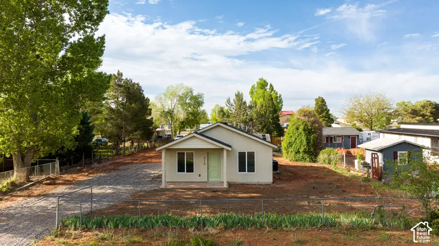 Single-story exterior featuring a light-toned stucco facade, covered front porch, gravel driveway, and detached storage structure situated on a fenced lot