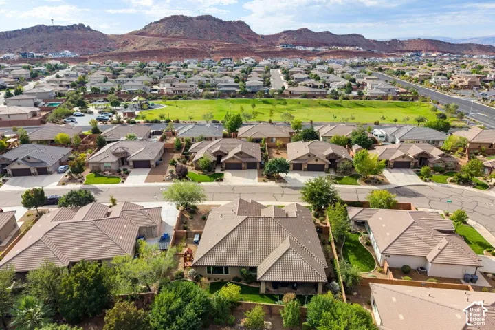 Aerial perspective of a residence featuring a multi-pitched tile roof, attached multi-car garage, fenced backyard with mature landscaping, and proximity to expansive open green space against a red rock mountain backdrop