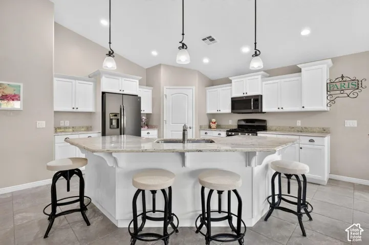 Kitchen featuring white shaker-style cabinetry, stone countertops, a center island with a built-in sink, stainless steel appliances, and suspended pendant lighting