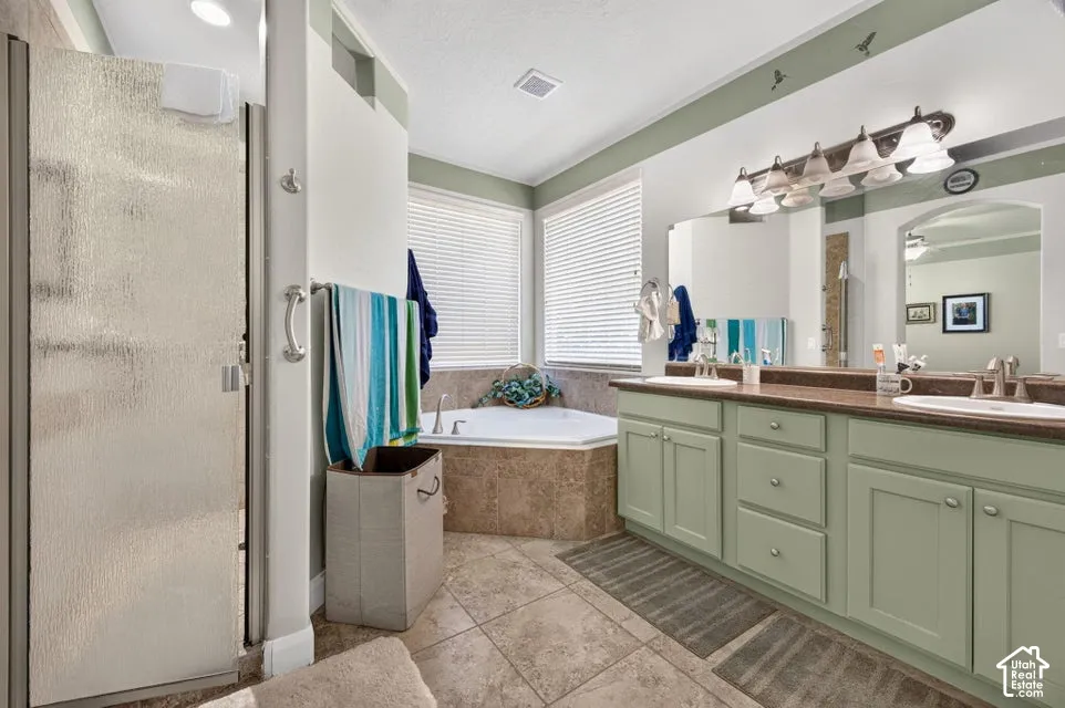 Dual-sink vanity with stone countertop, soaking tub with tile surround, glass-enclosed shower, ceramic tile flooring, and integrated wall-mounted mirror