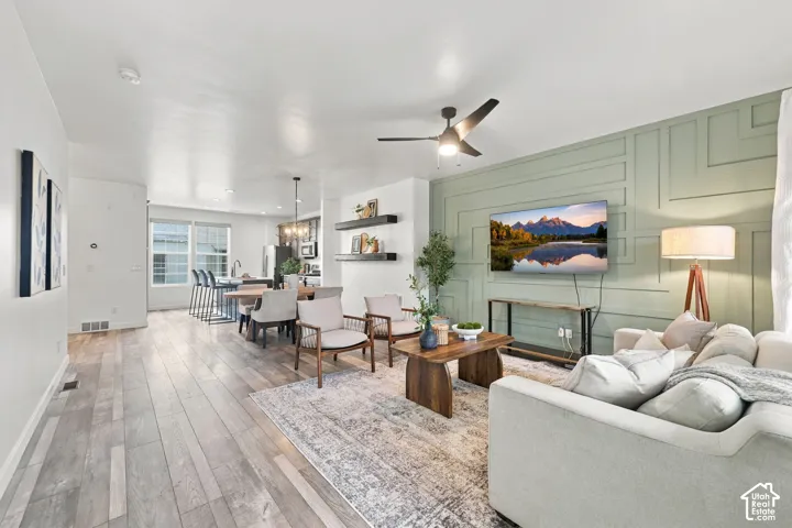 Open-concept living area with wood-finish flooring, a contemporary ceiling fan, and a light green accent wall with decorative paneling