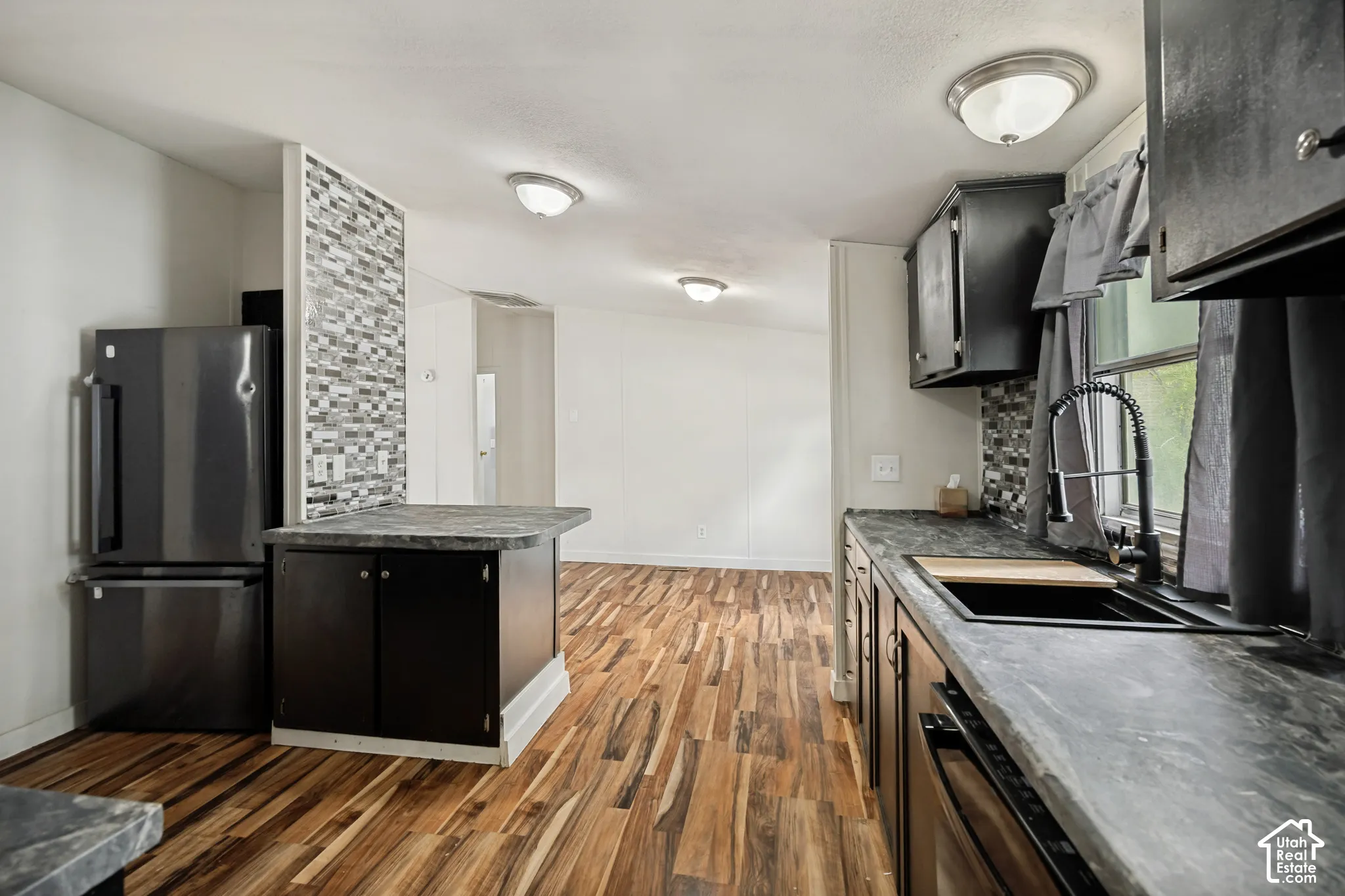 Kitchen featuring dark wood-finish cabinetry, a mosaic tile backsplash, black countertops, wood-finish flooring, and a stainless steel refrigerator