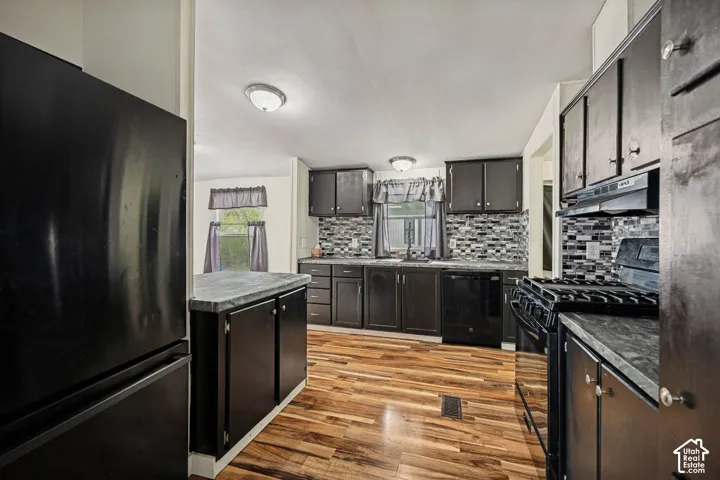 Black cabinetry with dark countertops, wood-finish flooring, mosaic tile backsplash, and stainless steel sink