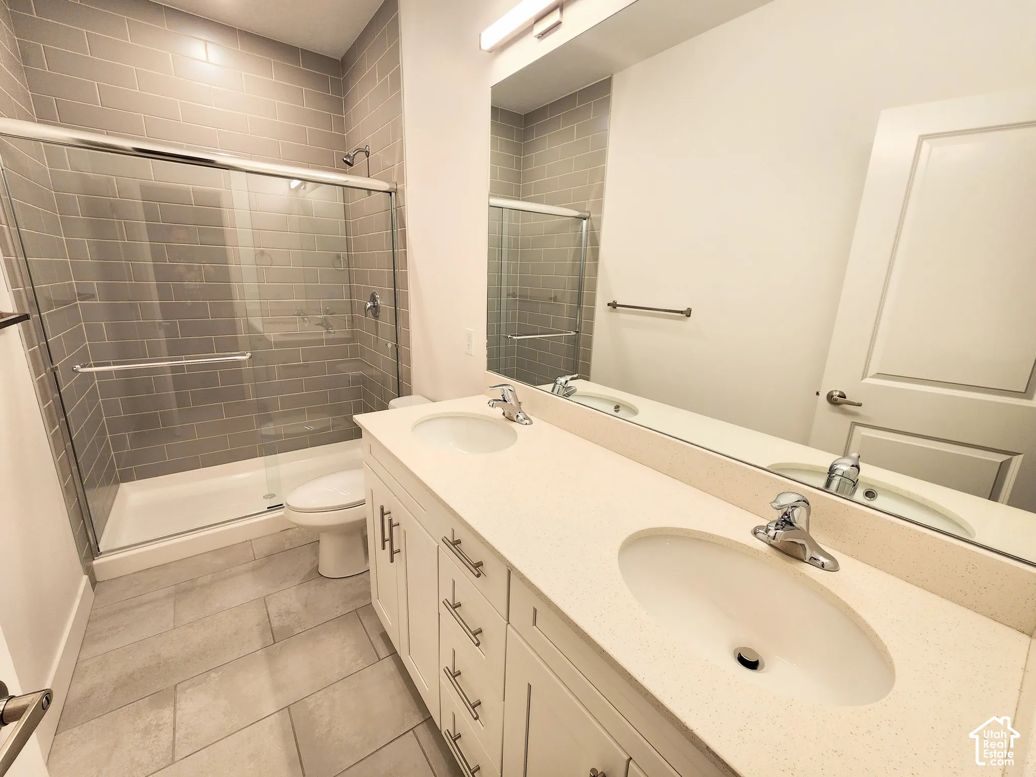 Double vanity featuring a light-toned solid surface countertop with dual undermount sinks, white cabinetry with metallic hardware, oversized frameless mirror, and a walk-in shower with grey subway tile surround