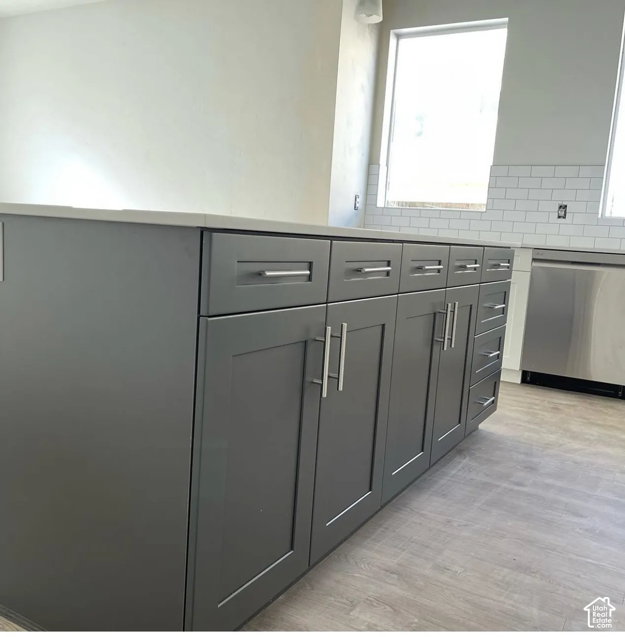 Kitchen island featuring slate-gray shaker-style cabinetry, brushed metal pulls, white countertop, white subway tile backsplash, and stainless steel dishwasher