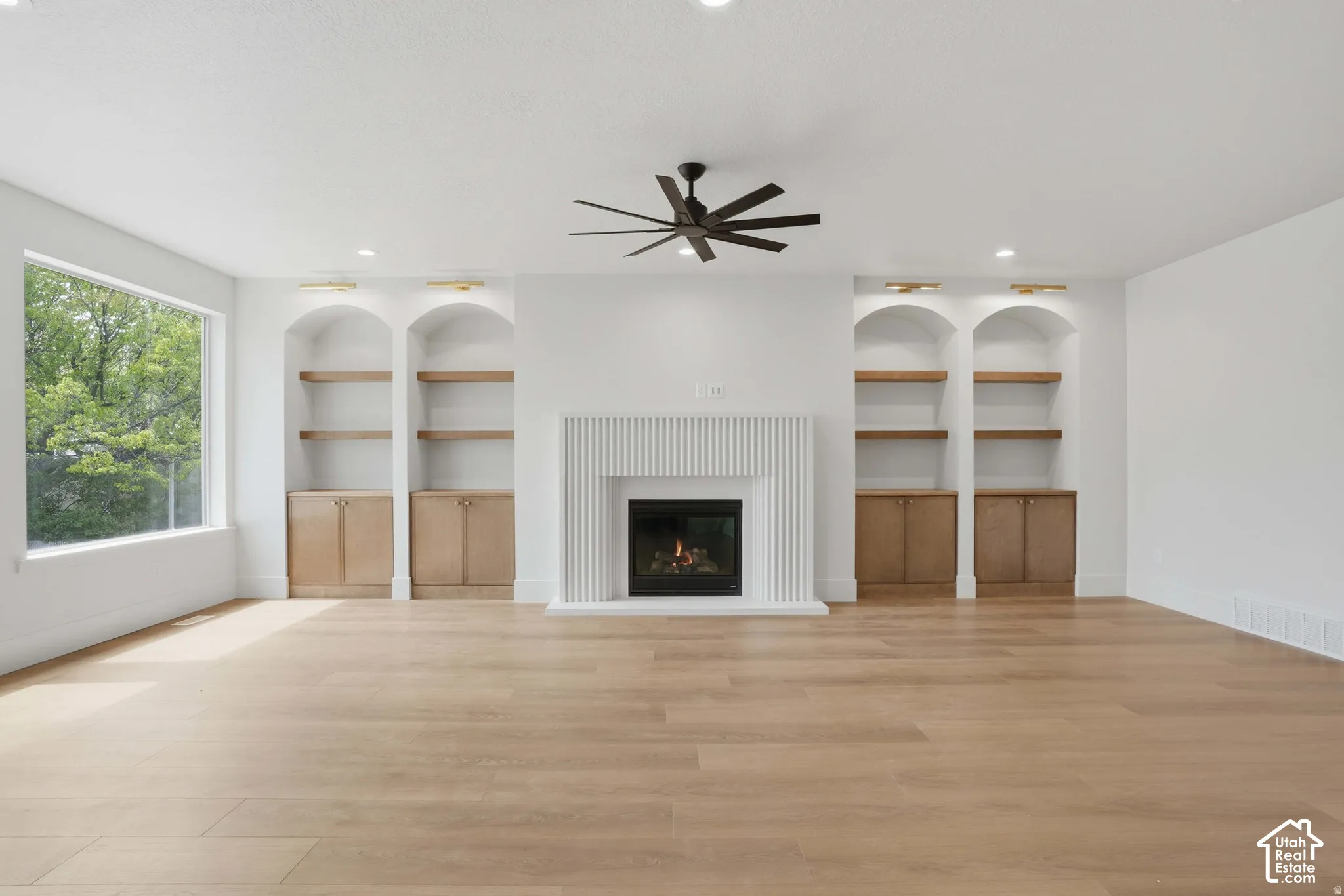 Fireplace with vertical fluted surround, four built-in arched alcoves with wood shelving and lower cabinetry, wood-finish flooring, and a matte black ceiling fan
