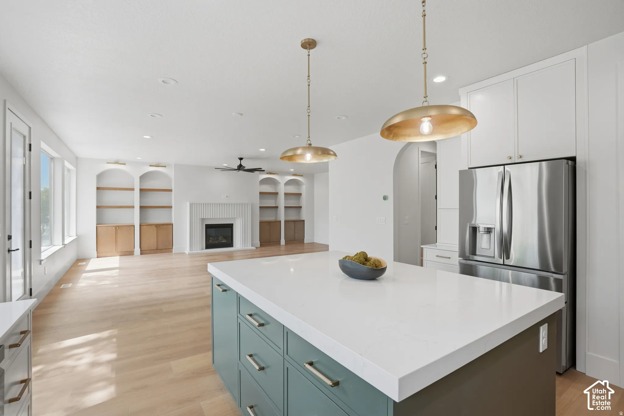 Kitchen island with a solid-surface countertop and integrated cabinetry, wood-finish flooring, dual metal pendant lighting, built-in recessed shelving, and a gas fireplace with a decorative mantel
