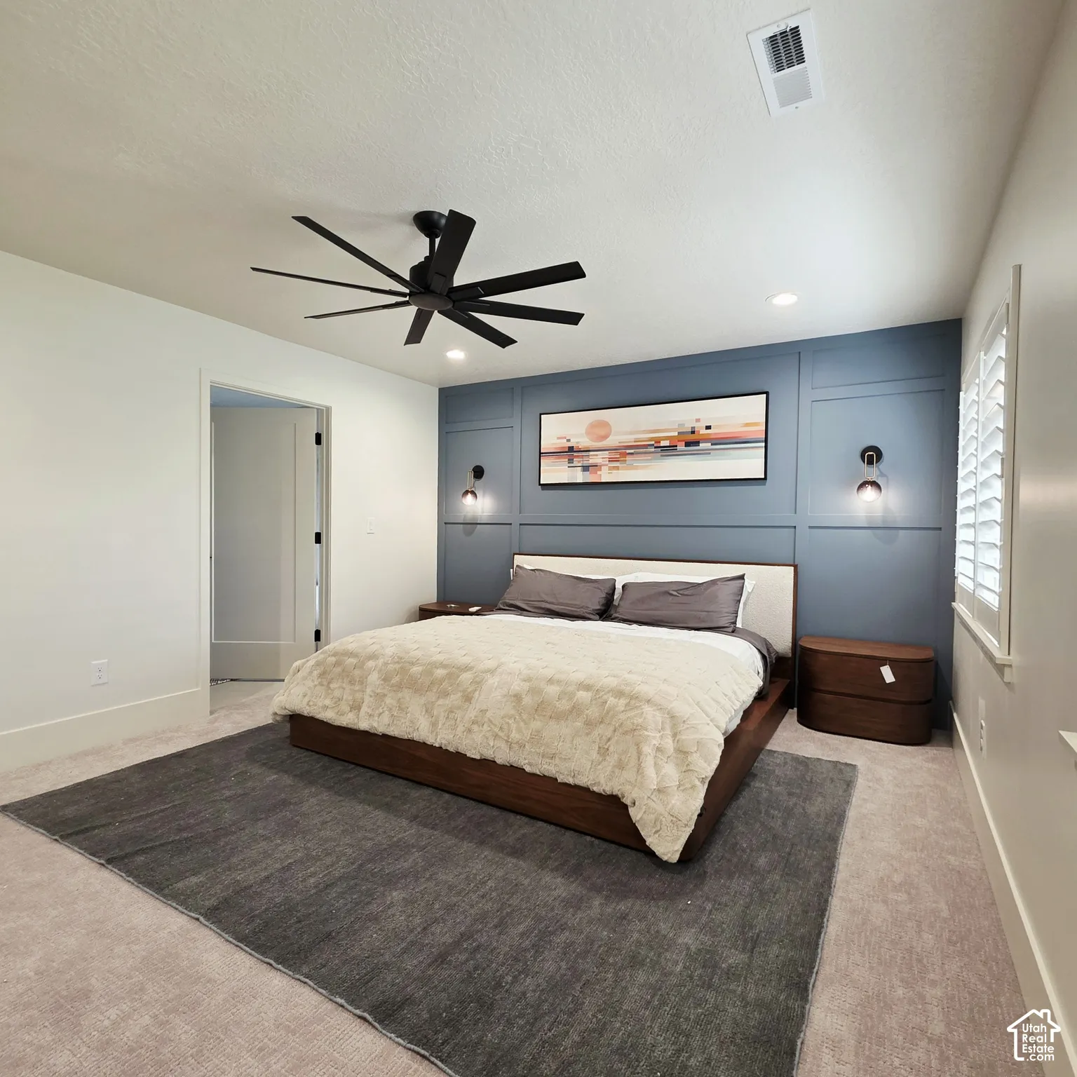 Blue panel accent wall, multi-blade matte black ceiling fan, dual wall-mounted light fixtures, and white plantation shutters