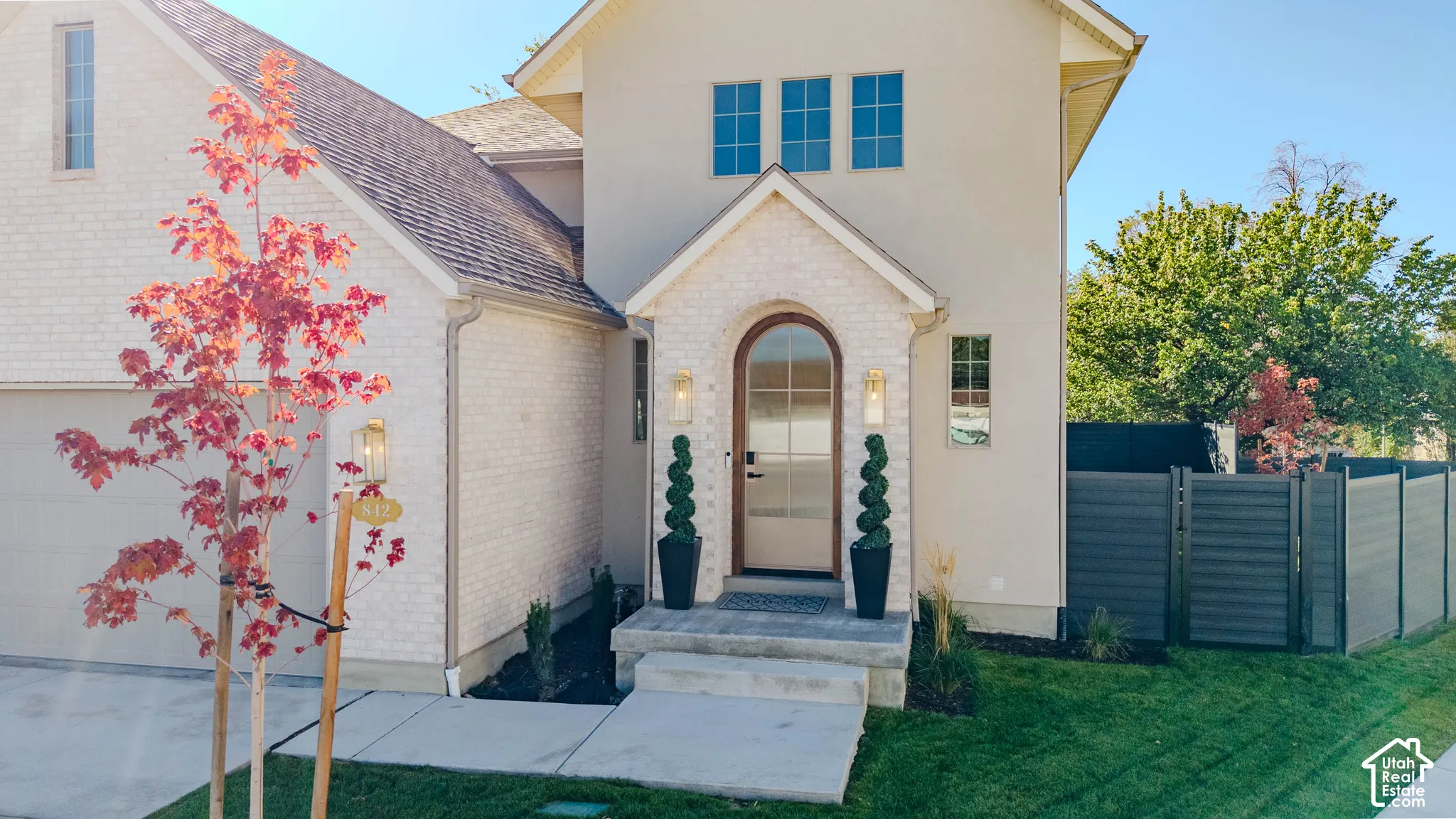 Exterior featuring light brick and stucco facade, arched entryway with glass-paneled door, metal wall-mounted lanterns, and horizontal privacy fencing