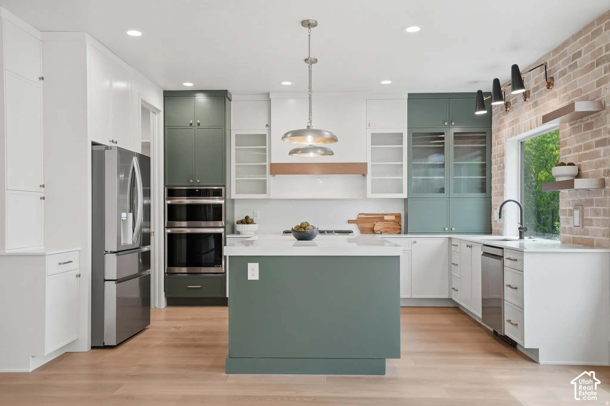Kitchen featuring two-tone cabinetry, central island with solid-surface countertop, stainless steel double wall ovens, exposed brick wall accent, and wood-finish flooring