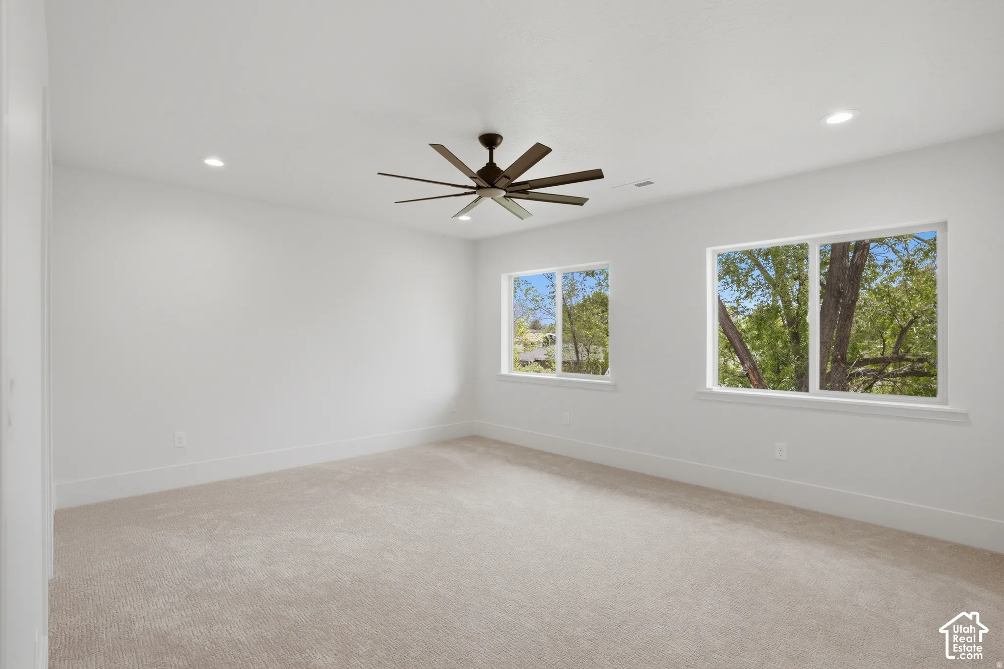 Neutral interior space featuring carpeting, recessed lighting, a multi-blade ceiling fan, and large windows with exterior foliage