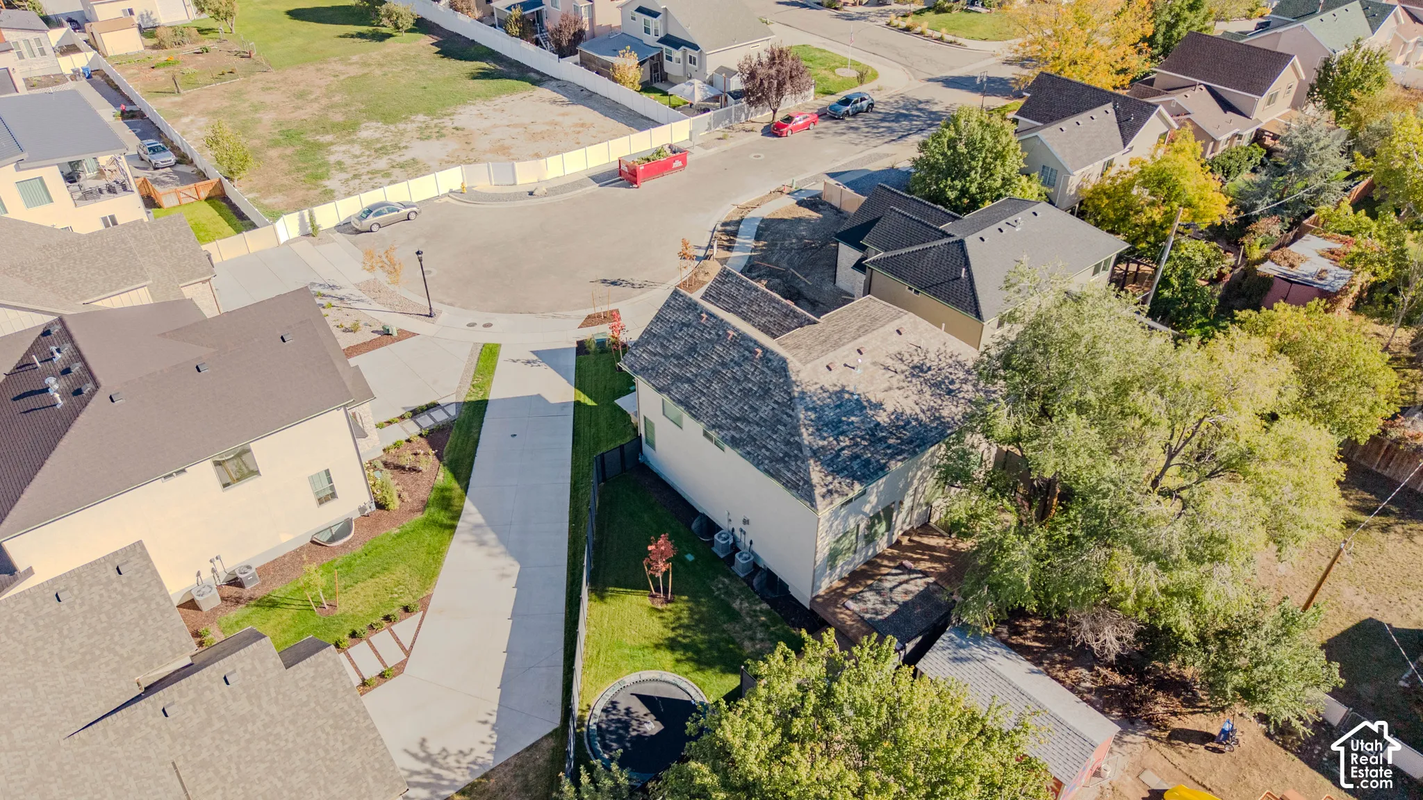 Aerial view of a residential property featuring a multi-level asphalt shingle roof, a private driveway, a fenced-in grass yard, and established trees