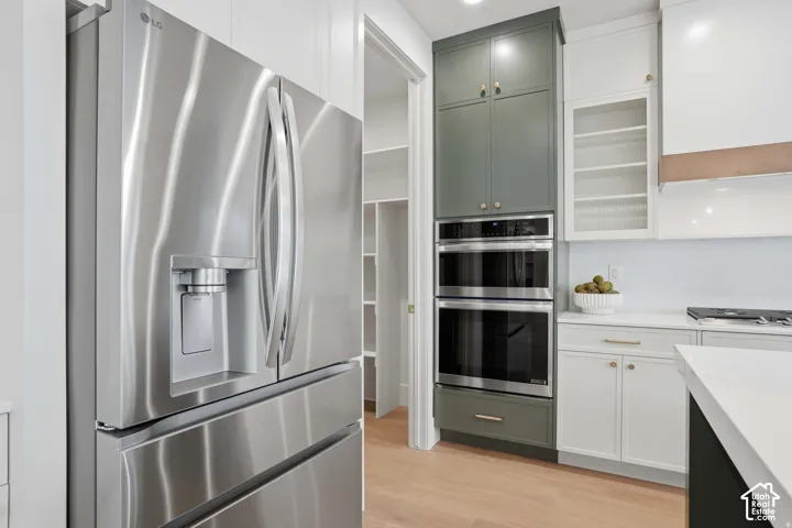 Stainless steel French-door refrigerator, double wall ovens, two-tone cabinetry with upper glass-front shelving, and wood-finish flooring