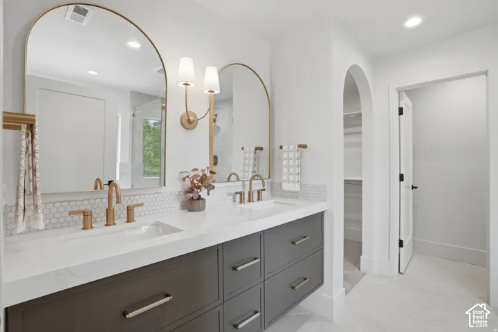 Dual vanity featuring quartz countertops, undermount sinks, brass-toned fixtures, arched mirrors, and grey shaker-style cabinetry with herringbone-patterned tile flooring