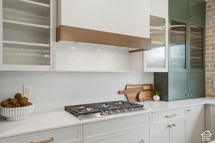 Kitchen featuring a stainless steel gas cooktop, custom hood with wood-finish trim, slab stone countertops, contrasting cabinetry with glass-panel doors, and an exposed brick wall