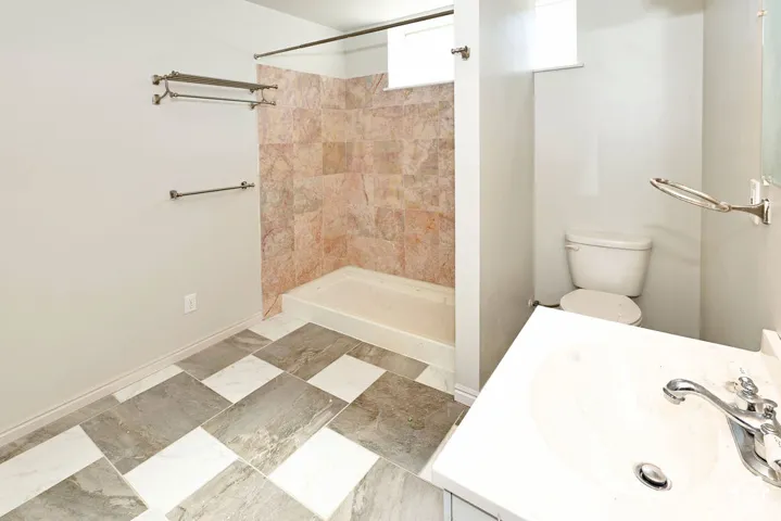 Tiled shower surround, white shower base, ceramic commode, white vanity with integrated sink, and checkerboard floor tiles