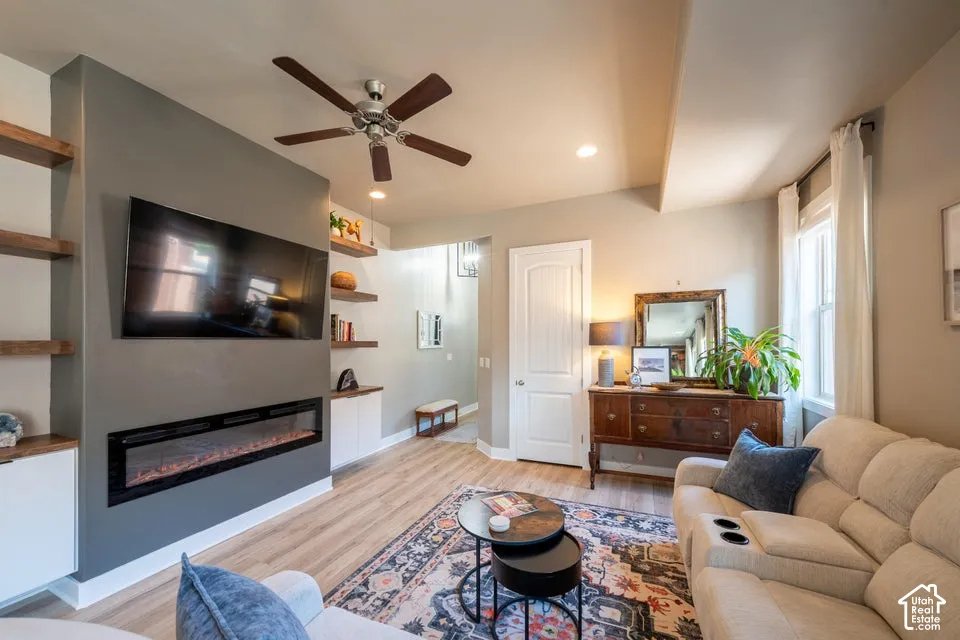 Living space featuring wood-finish flooring, a linear electric fireplace, custom built-in cabinetry, wood-finish wall shelving, and a ceiling fan