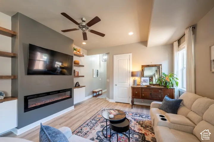 Living space featuring wood-finish flooring, a linear electric fireplace, custom built-in cabinetry, wood-finish wall shelving, and a ceiling fan