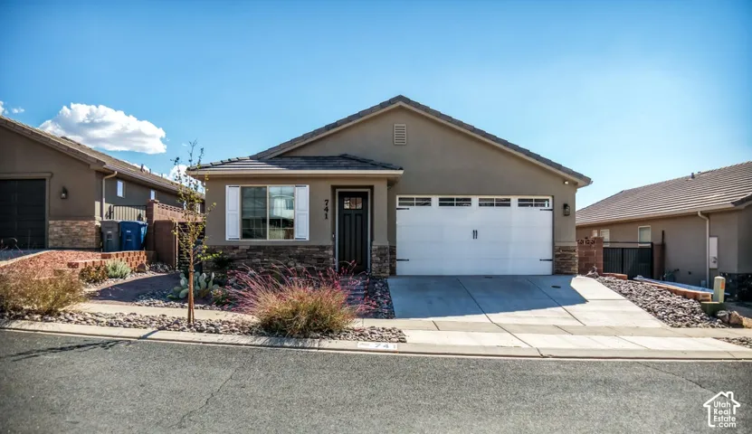 Single-story exterior featuring a stucco and stone veneer facade, gabled roofline, two-car garage with windows, and low-maintenance xeriscaped front yard