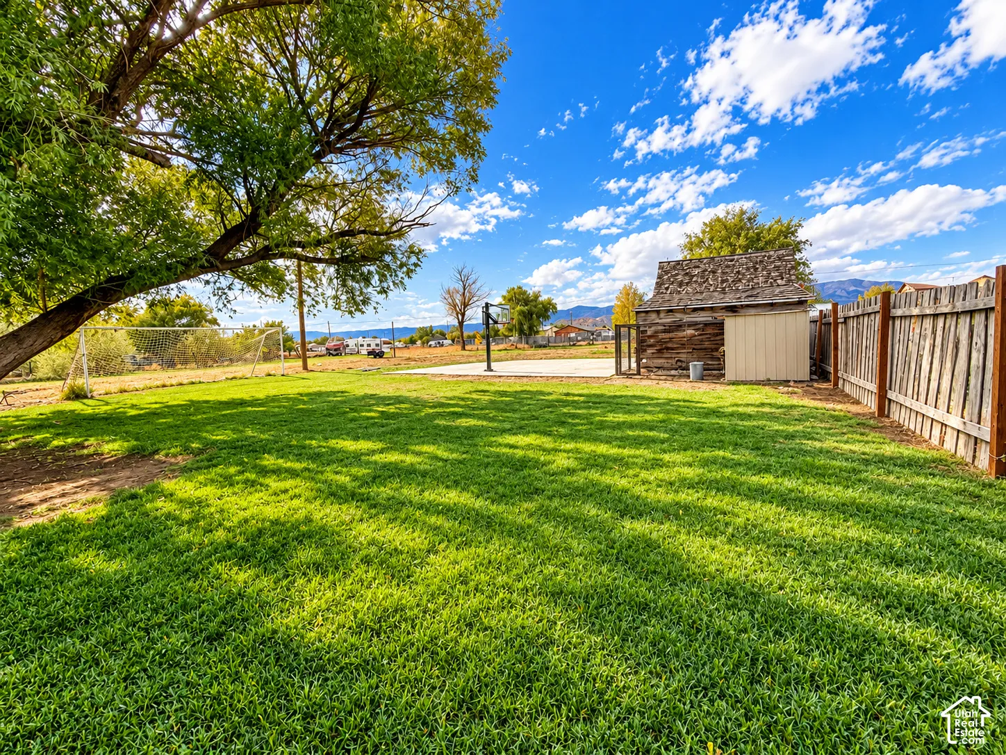 Expansive backyard featuring a grass lawn, mature trees, wood fence, concrete pad, and a wooden storage structure
