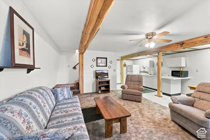 Open-concept interior featuring exposed structural wood beams, ceiling-mounted paddle fan, and kitchen area with white cabinetry