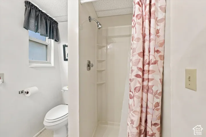 Bathroom featuring a white elongated toilet, a built-in shower stall with integrated shelving, a chrome-finish shower head, and a single-pane window