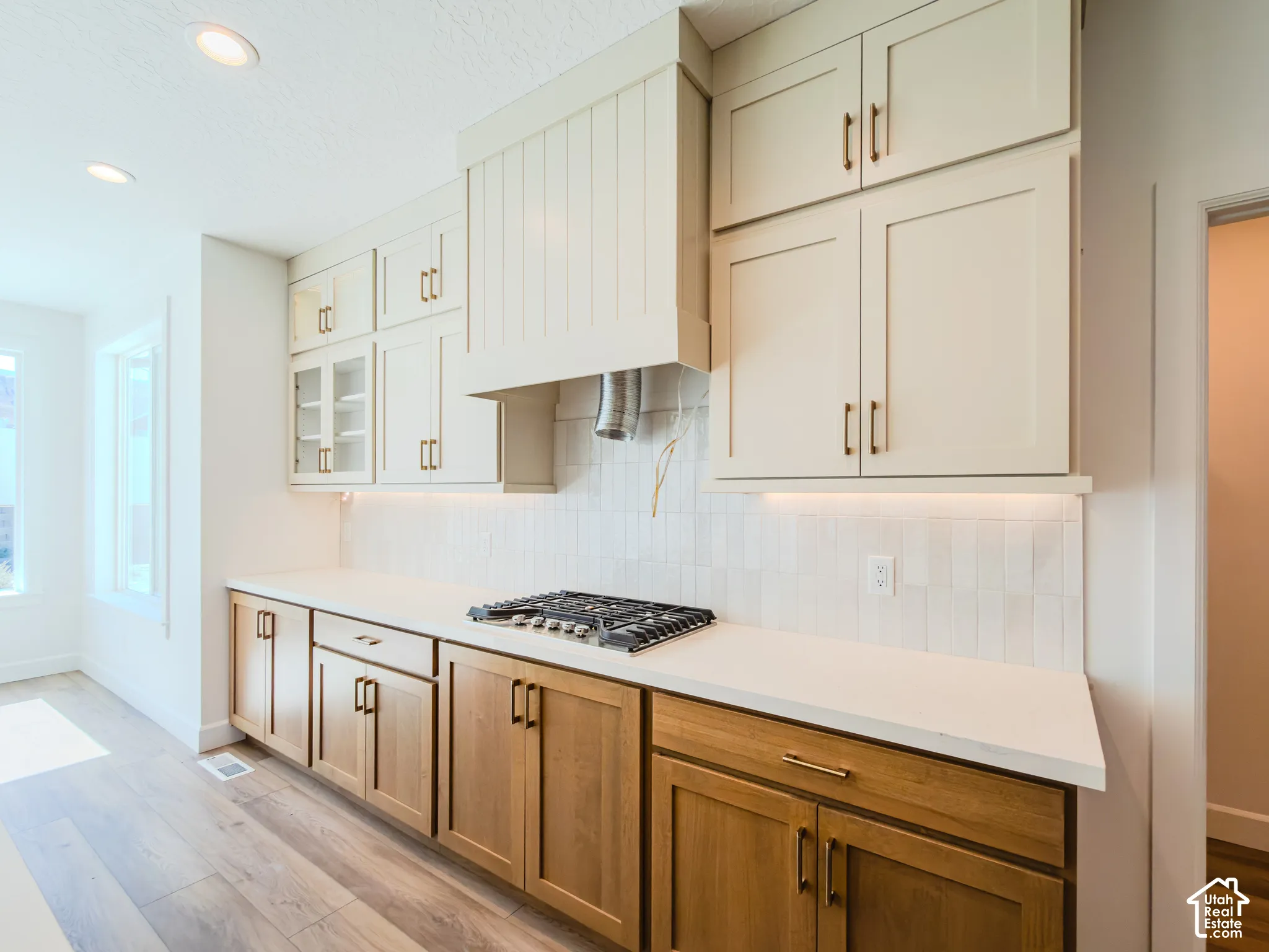 Kitchen featuring two-tone cabinetry with upper shaker-style doors, natural wood-finish lower cabinetry, solid-surface countertops, textured tile backsplash, and a built-in cooktop