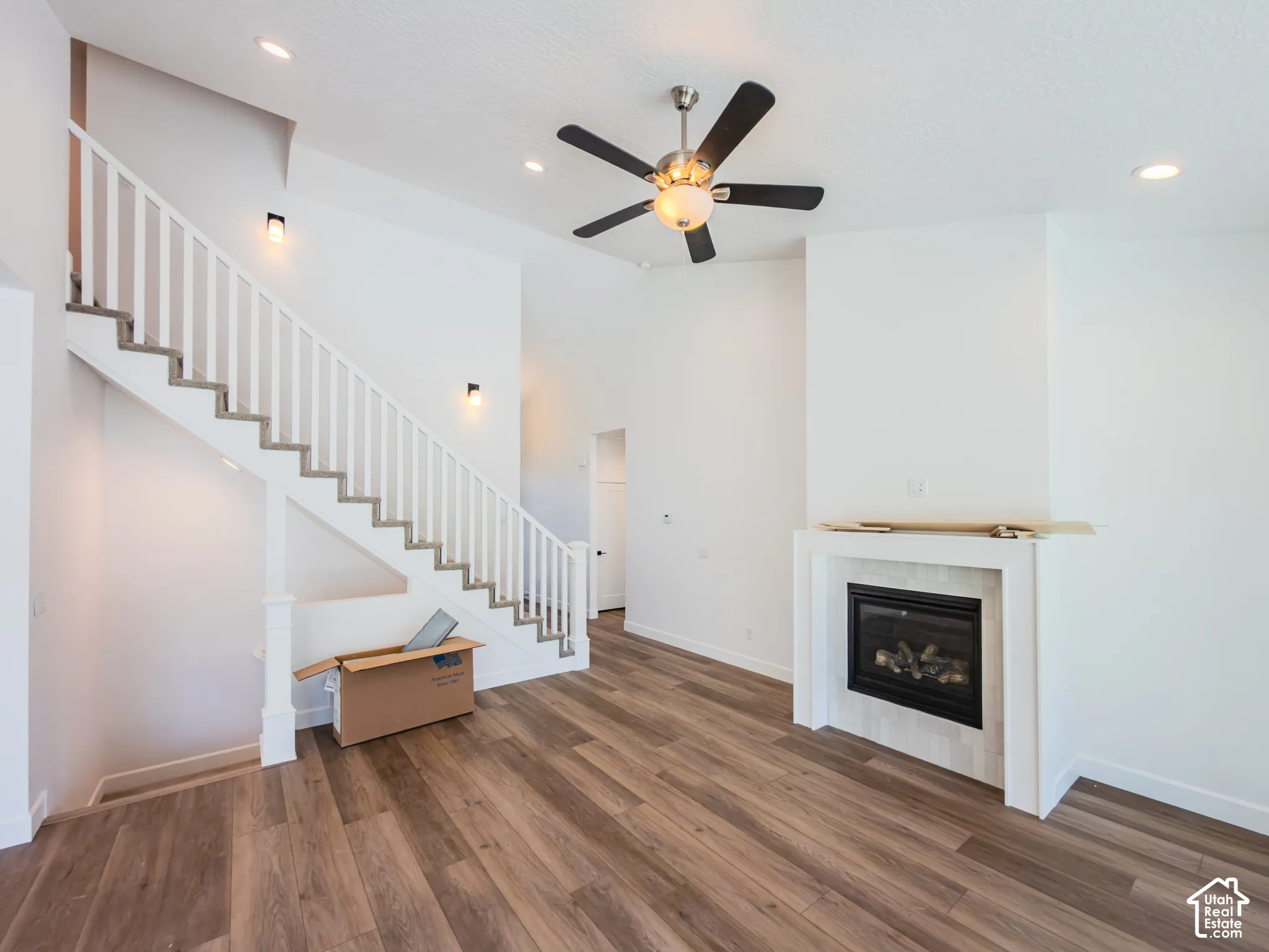 High-ceiling interior featuring wood-finish flooring, a fireplace with a light-toned surround, a staircase with white balusters, recessed lighting, and a ceiling fan