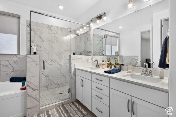 Dual-sink vanity with white cabinetry and solid surface countertop, walk-in shower with floor-to-ceiling stone-finish tile surround and frameless glass enclosure, and a built-in soaking tub