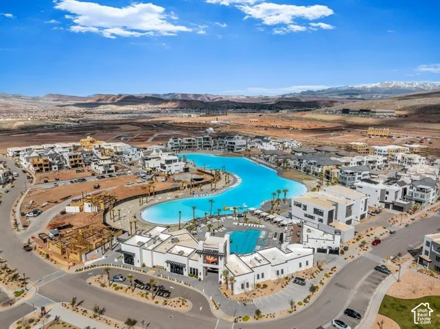 Aerial perspective of a multi-unit residential community featuring a large, central turquoise recreational lagoon, a swimming pool, and a clubhouse facility, set against a desert landscape with mountain backdrops
