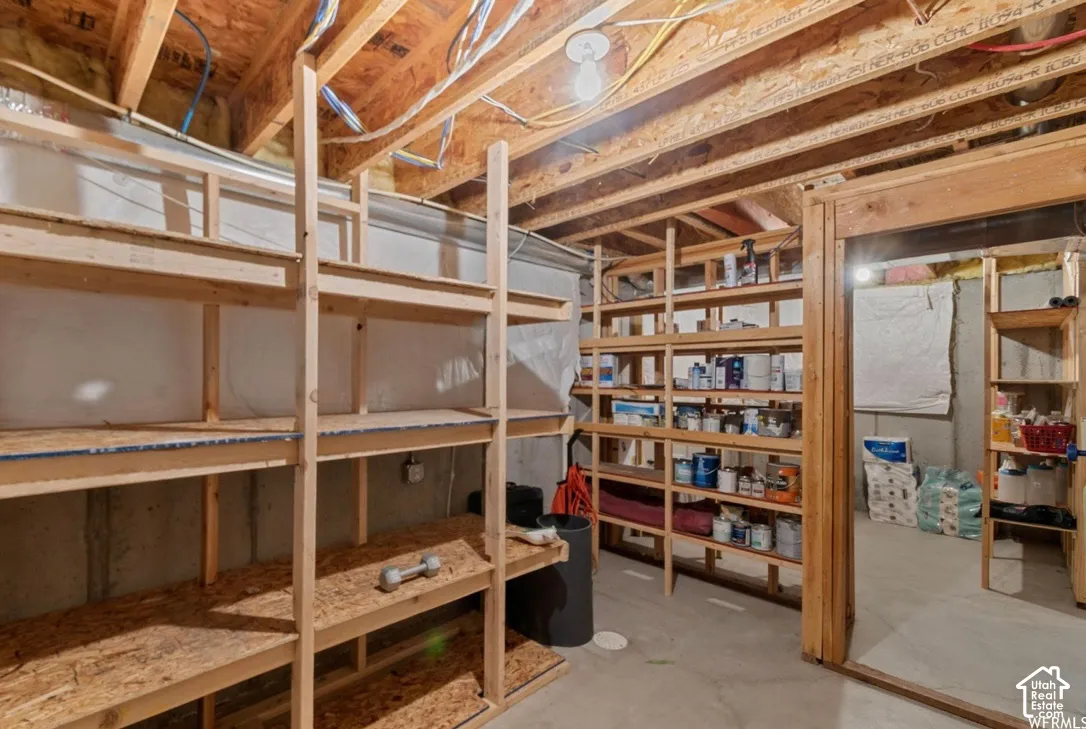 Unfinished storage area featuring concrete foundation walls, exposed wood ceiling joists, poured concrete flooring, and extensive built-in timber shelving