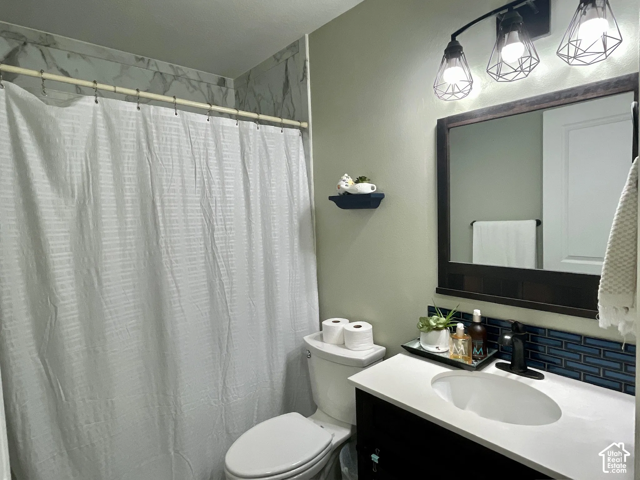 Bathroom featuring a vanity with a white countertop and oval under-mount sink, dark tile backsplash, three-light geometric wall fixture, and shower enclosure with decorative wall tile