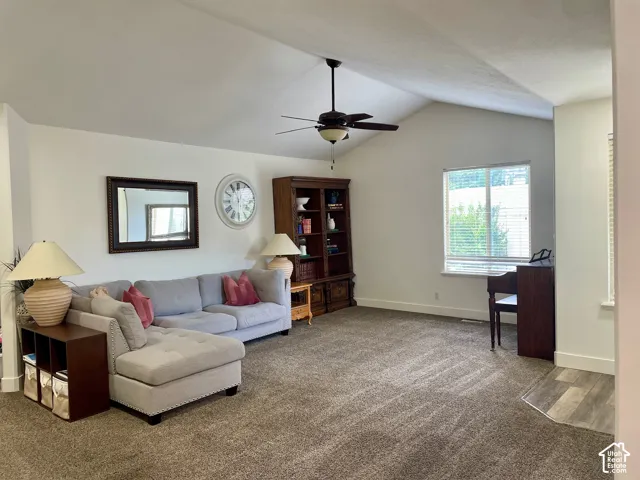 Living area featuring vaulted ceilings, neutral wall tones, wall-to-wall carpeting, a ceiling fan with integrated lighting, and a hearth finished with wood-finish flooring